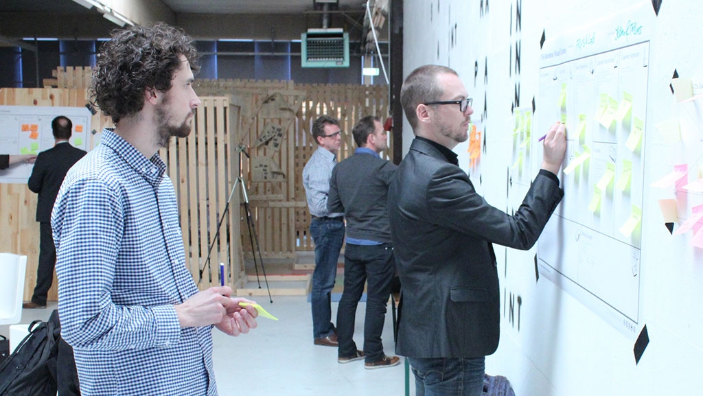 Das Bild zeigt vier Personen an einer Planwand. Eine Person schreibt eine Notiz auf einen Haftzettel an der Wand und wird dabei von einer weiteren Person beobachtet.